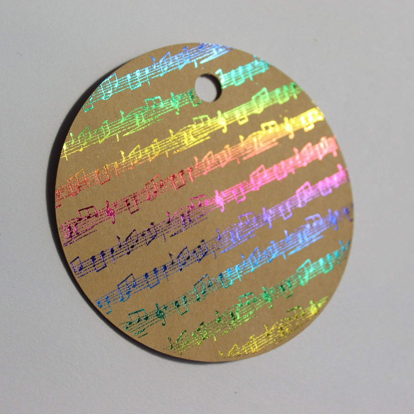 Round brown kraft gift tag. Design of rows of music in a multicoloured shiny foil.