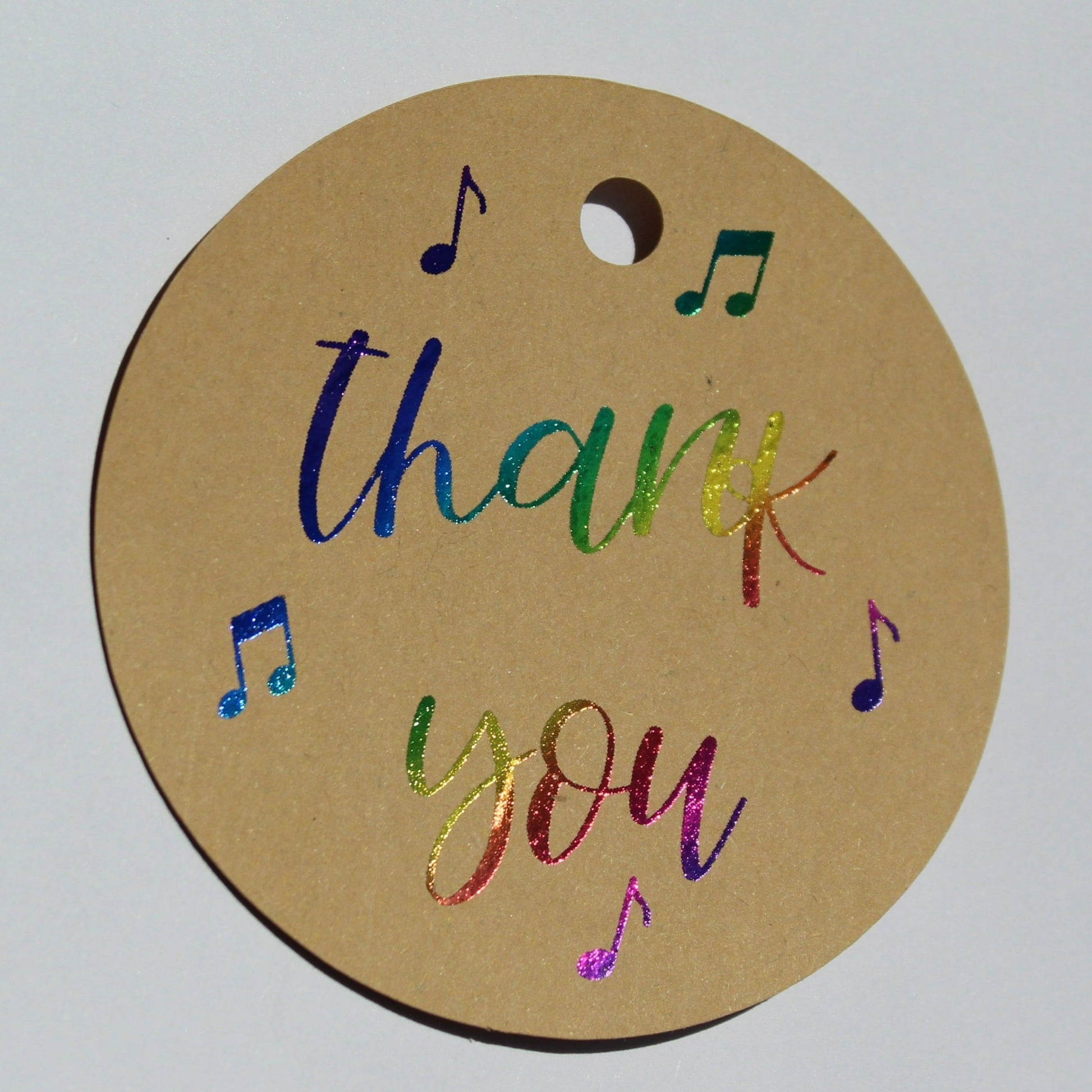 Round brown kraft gift tag. Text reads 'thank you' in multicoloured shiny foil with several music notes around the text.