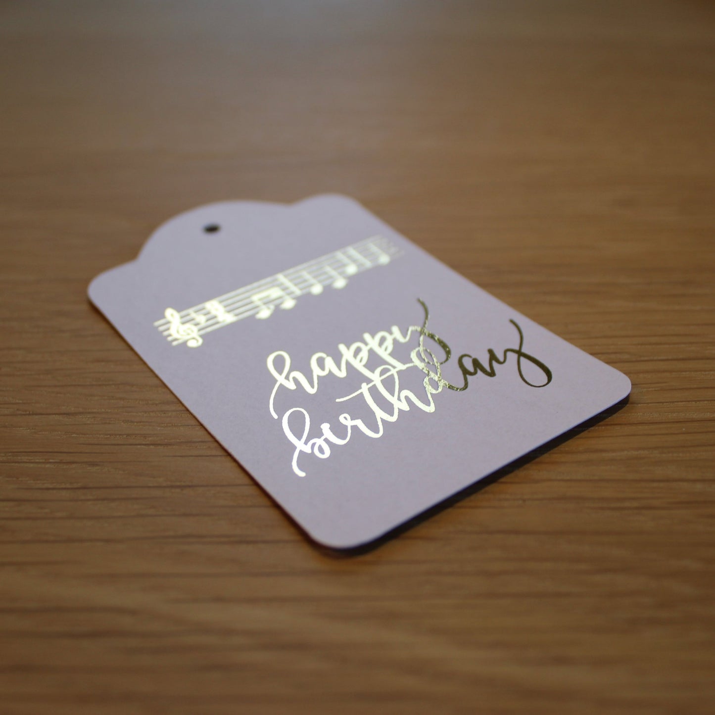 Oblong white tag with 'happy birthday' and the music of 'happy birthday to you' displayed in gold shiny foil.